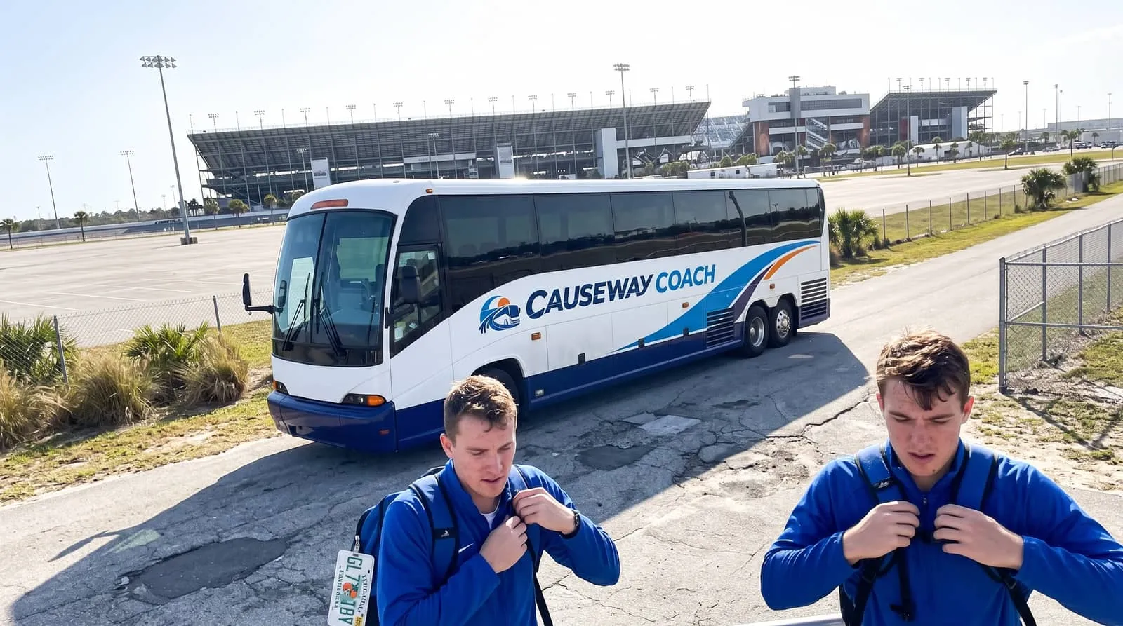 University athletes with kit bags beside a Causeway Coach motorcoach on a service road outside the Daytona Beach speedway