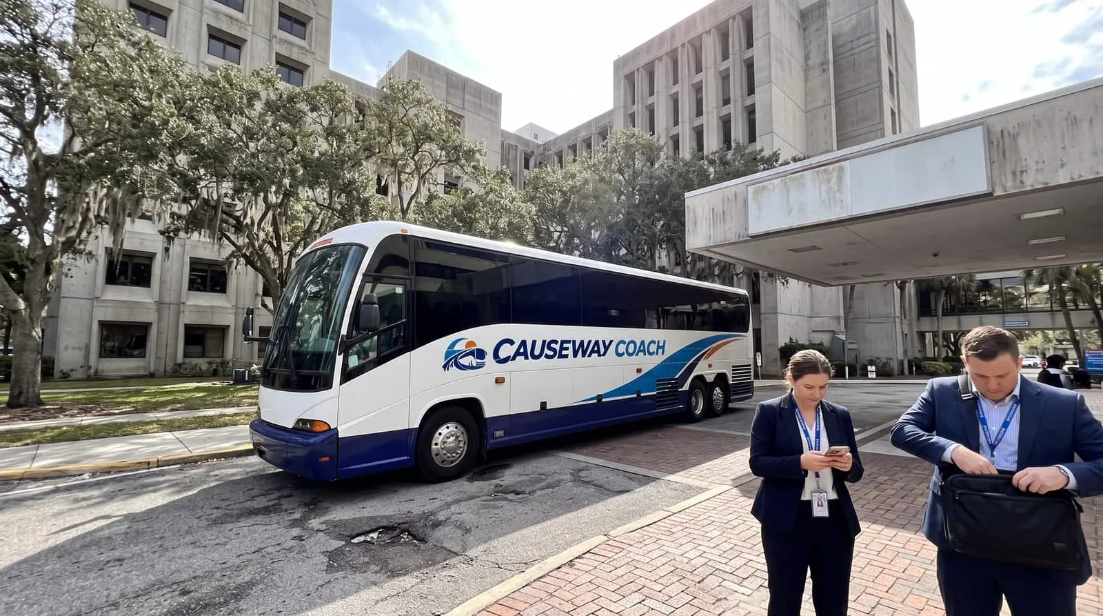 Two people with lanyards beside a Causeway Coach motorcoach at the entrance drive of a Tampa medical campus